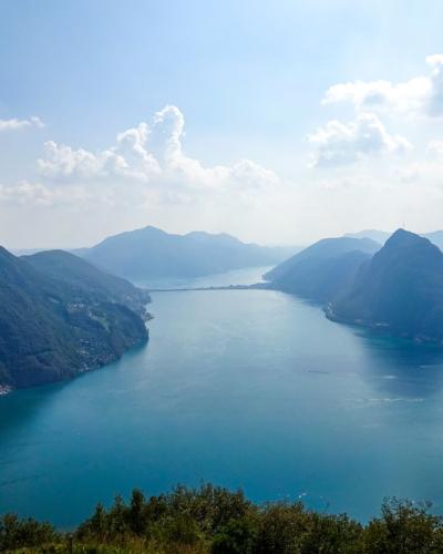 Breathtaking aerial view of Lake Lugano surrounded by lush mountains and clear blue skies.